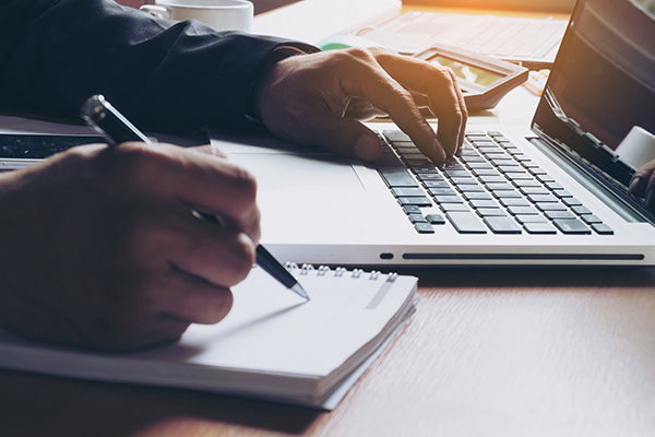 Asset-Liability-Services_V2 closeup of person writing on paper with a pen, while typing on a laptop keyboard
